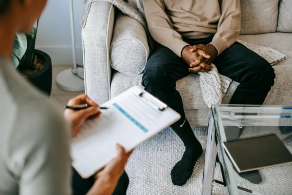 A consultant holding a clipboard while speaking with a client seated on a couch
